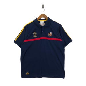 May include: Navy blue polo shirt with yellow stripes on the sleeves and collar. A red horizontal stripe crosses the chest. Features the Adidas logo and a World Cup emblem. The shirt is hanging on a wooden hanger.