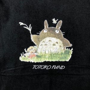 May include: Black sweatshirt featuring a graphic of Totoro, a large, friendly forest spirit, with a young girl in a field of green grass. The text "TOTORO FUND" is printed in white below the image. The design is a whimsical illustration.