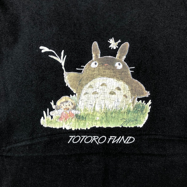 May include: Black sweatshirt featuring a graphic of Totoro, a large, friendly forest spirit, with a young girl in a field of green grass. The text "TOTORO FUND" is printed in white below the image. The design is a whimsical illustration.