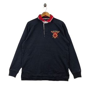 Puede incluir: Una camiseta de rugby de manga larga azul marino con cuello rojo y blanco. La camiseta presenta un escudo bordado con el texto "Hilfiger" y un diseño de remo.