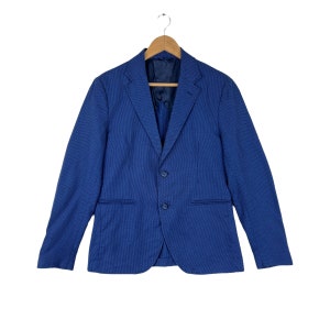 Peut inclure: Un blazer bleu avec une fermeture à deux boutons et des revers crantés est suspendu à un cintre en bois. Le blazer a deux poches avant et une doublure bleu foncé. Le tissu présente un motif texturé subtil.