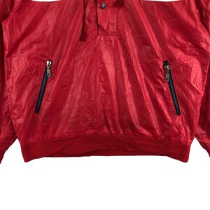 May include: A red nylon jacket with a quarter-zip closure and two zippered pockets. The jacket has a ribbed hem and cuffs. The brand logo is visible on the zipper pulls. The jacket is a lightweight, sporty style.