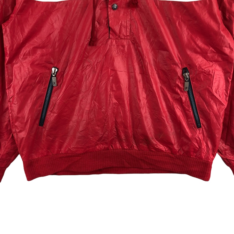 May include: A red nylon jacket with a quarter-zip closure and two zippered pockets. The jacket has a ribbed hem and cuffs. The brand logo is visible on the zipper pulls. The jacket is a lightweight, sporty style.