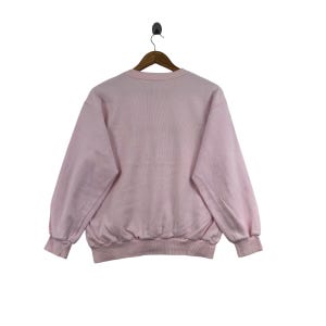 May include: A light pink sweatshirt with a crew neck and long sleeves. The sweatshirt is hanging on a wooden hanger against a white background. The cuffs and hem are ribbed.