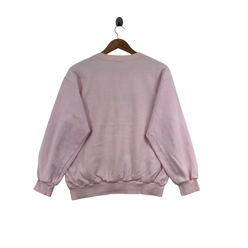 May include: A light pink sweatshirt with a crew neck and long sleeves. The sweatshirt is hanging on a wooden hanger against a white background. The cuffs and hem are ribbed.