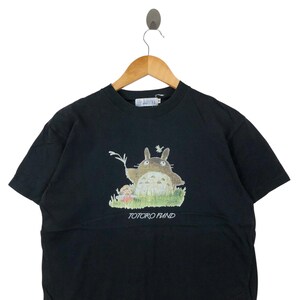 May include: Black short-sleeved t-shirt featuring a graphic of Totoro from the animated film My Neighbor Totoro. The image shows Totoro in a grassy field with a small character. The text "TOTORO FUND" is printed below the graphic.