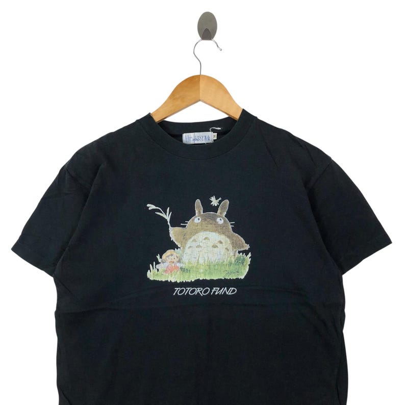 May include: Black short-sleeved t-shirt featuring a graphic of Totoro from the animated film My Neighbor Totoro. The image shows Totoro in a grassy field with a small character. The text "TOTORO FUND" is printed below the graphic.