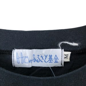 May include: Close-up of a black crew neck shirt with a white tag. The tag features Japanese text in blue and a size "M" label. The shirt's fabric appears to be a soft, knit material.