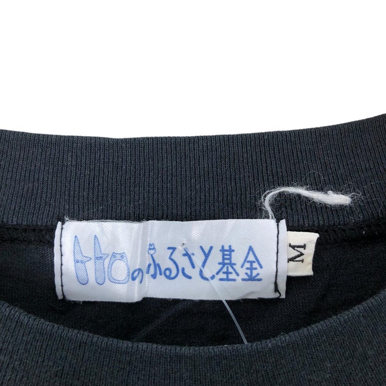 May include: Close-up of a black crew neck shirt with a white tag. The tag features Japanese text in blue and a size "M" label. The shirt's fabric appears to be a soft, knit material.