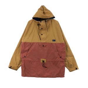 May include: A tan and rust anorak jacket with a hood. The jacket has a half-zip, a front pocket, and a drawstring waist. It features a small logo patch and is made of lightweight material. The jacket is suitable for outdoor activities.