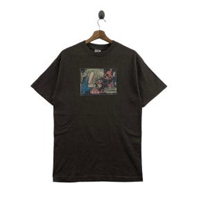 Puede incluir: Camiseta gris oscuro con estampado de panel de cómic. El gráfico muestra una escena con figuras en acción, incluyendo una mujer rubia con un vestido azul y una figura enmascarada de rojo. La camiseta está colgada de una percha de madera.