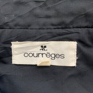 K&ouml;nnte beinhalten: Ein wei&szlig;es Etikett mit schwarzem Text, der "Courr&egrave;ges" lautet, mit einem kleinen "CC"-Logo &uuml;ber dem Text. Das Etikett ist an einem schwarzen Stoff befestigt.