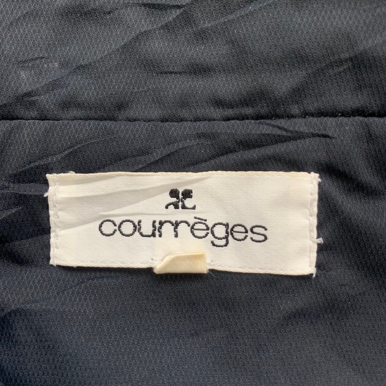 K&ouml;nnte beinhalten: Ein wei&szlig;es Etikett mit schwarzem Text, der "Courr&egrave;ges" lautet, mit einem kleinen "CC"-Logo &uuml;ber dem Text. Das Etikett ist an einem schwarzen Stoff befestigt.
