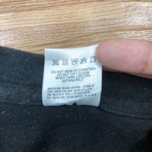 May include: White care label with black text on a black t-shirt. The label has instructions for washing and drying the garment. The text reads: "DO NOT IRON DECORATION DO NOT DRY CLEAN WASH DARK COLORS SEPARATELY MACHINE WASH WARM, TUMBLE DRY MEDIUM USE NON-CHLORINE BLEACH ONLY IF NEEDED. CA 18345 RN 13785 MADE IN USA"