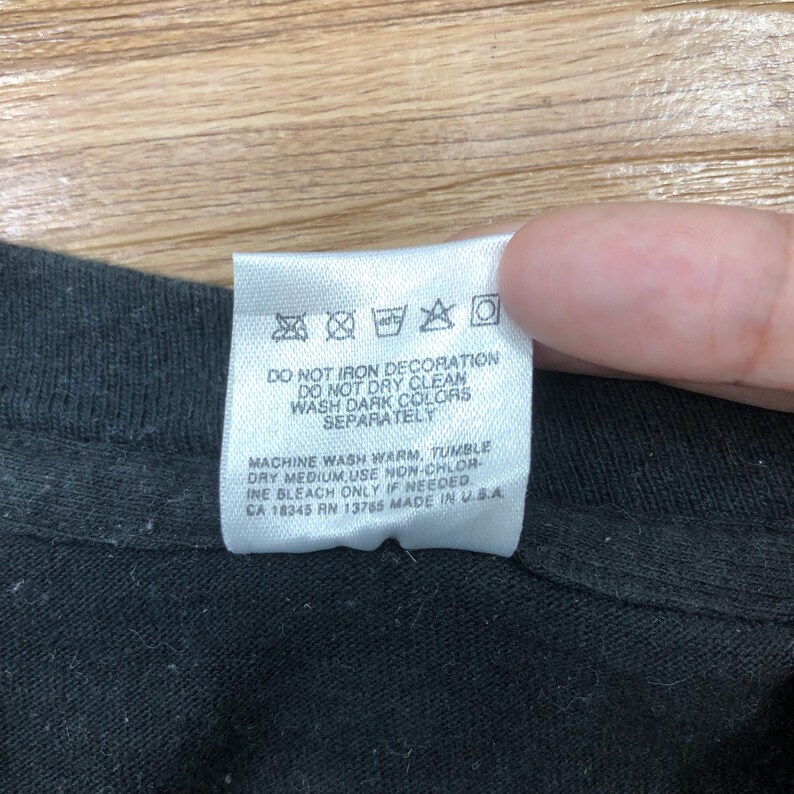 May include: White care label with black text on a black t-shirt. The label has instructions for washing and drying the garment. The text reads: "DO NOT IRON DECORATION DO NOT DRY CLEAN WASH DARK COLORS SEPARATELY MACHINE WASH WARM, TUMBLE DRY MEDIUM USE NON-CHLORINE BLEACH ONLY IF NEEDED. CA 18345 RN 13785 MADE IN USA"