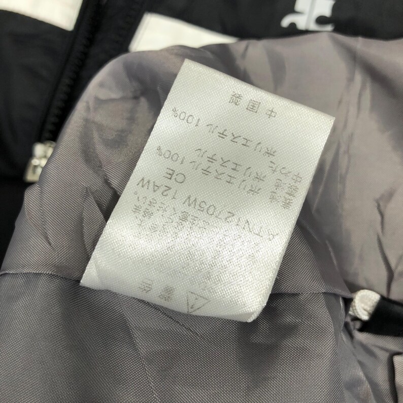 May include: Close-up of a white tag with Japanese text and the text "ATN12705W 12AW" on a gray lining. The tag also includes the text "CE" and "ポリエステル 100%" and a warning symbol. The background is black and white.