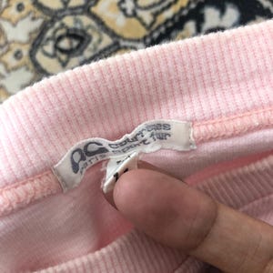 May include: Close-up of a pink ribbed fabric garment with a tag. The tag is white with black text and the letters "ac paris sport fur". The garment is a light pink color.