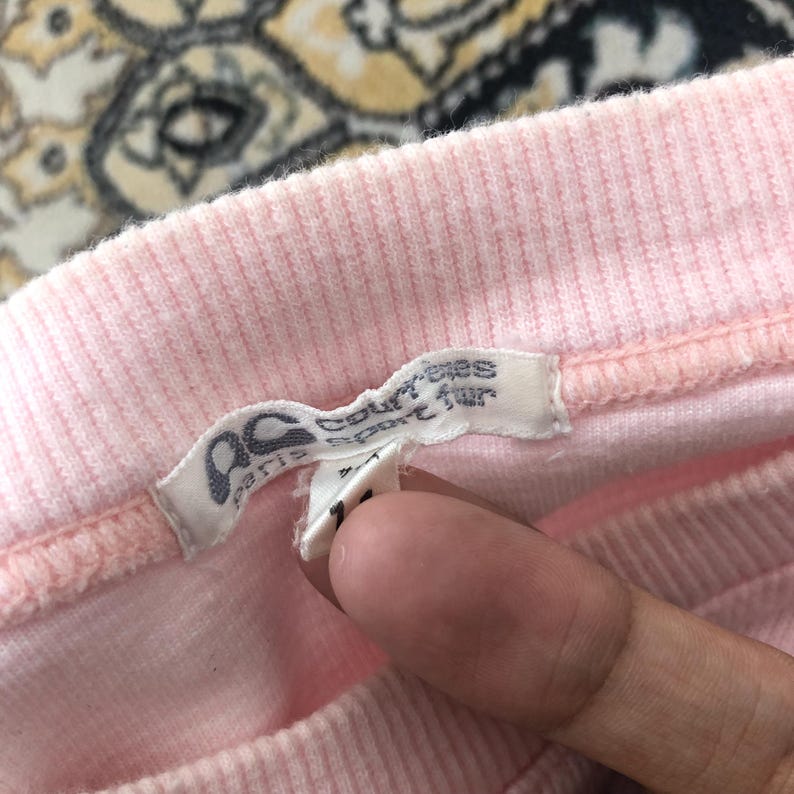 May include: Close-up of a pink ribbed fabric garment with a tag. The tag is white with black text and the letters "ac paris sport fur". The garment is a light pink color.