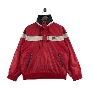 May include: Red vintage windbreaker with white and navy blue accents. Features a high collar, a button-up placket, and two zippered pockets. The Fila logo is on the chest. A classic athletic jacket.