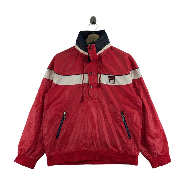 May include: Red vintage windbreaker with white and navy blue accents. Features a high collar, a button-up placket, and two zippered pockets. The Fila logo is on the chest. A classic athletic jacket.