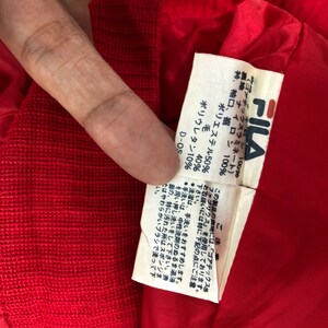 May include: Close-up of a red garment's interior, revealing a white tag with the FILA logo and Japanese text. The tag lists fabric composition: polyester 50%, wool 40%, and polyurethane 10%.