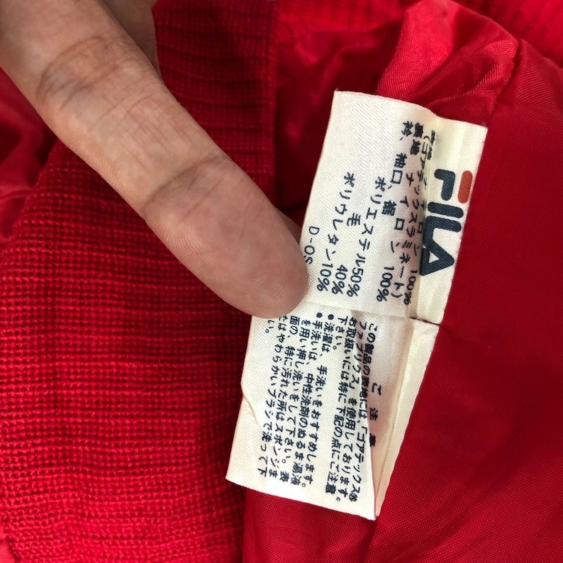 May include: Close-up of a red garment's interior, revealing a white tag with the FILA logo and Japanese text. The tag lists fabric composition: polyester 50%, wool 40%, and polyurethane 10%.