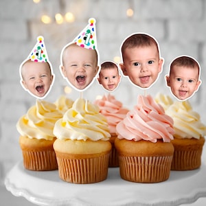 K&ouml;nnte beinhalten: Cupcakes mit gelbem und rosa Zuckerguss, belegt mit Babygesicht-Ausschnitten. Einige Cupcakes haben ein Baby mit einer Partyhut mit der Zahl 1. Die Cupcakes stehen auf einer wei&szlig;en Tortenplatte.