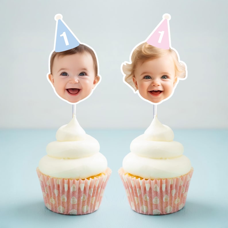 May include: Two cupcakes with white frosting and pink polka dot liners. Each cupcake has a photo of a baby's face on a stick, wearing a party hat with the number 1. One hat is blue, the other is pink.