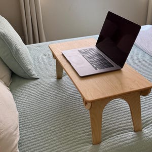 May include: A light wood laptop stand with a laptop on top of it. The stand is on a light blue bed with white and light blue pillows.