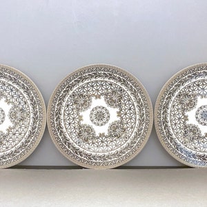 Könnte beinhalten: Drei dekorative Teller mit weißem Hintergrund und aufwendigen goldenen und silbernen Designs. Die Teller sind rund und haben einen erhöhten Rand.