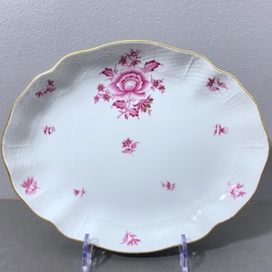 Könnte beinhalten: Weiße ovale Servierplatte mit goldfarbenem Rand und rosafarbenem Blumendesign. Das Design zeigt eine große rosafarbene Rose mit kleineren rosafarbenen Blüten und Blättern.