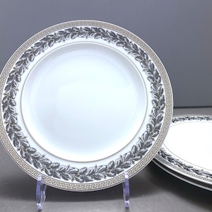 May include: A white porcelain dinner plate featuring a decorative border with a black and white laurel leaf wreath pattern, accented by a gold Greek key geometric design and a thin gold beaded rim detail along the inner edge of the border.