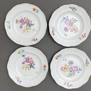 May include: Four white ceramic plates with scalloped edges and a textured rim. Each plate features a central floral bouquet in shades of pink, purple, yellow, and green. The plates are arranged on a gray surface.