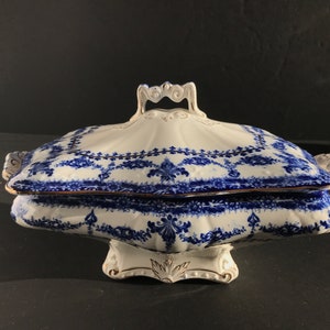 Może przedstawiać: Biała porcelanowa miska do serwowania z niebieskim i złotym wzorem kwiatowym. Miska ma pokrywkę z uchwytem i kwadratową podstawę.