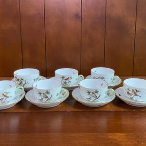 May include: A set of four white porcelain teacups and matching saucers, each featuring a textured basket-weave pattern on the exterior and a colorful illustration of small birds perched on branches. The cups have light green handles and gold rims.