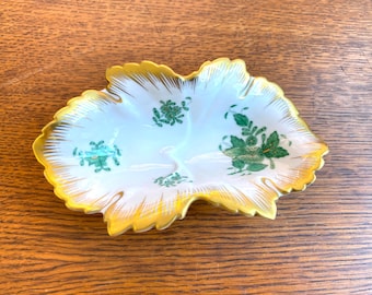May include: A white porcelain dish shaped like a leaf with a gold rim and green floral designs.