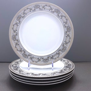 Puede incluir: Juego de cuatro platos de cena de cerámica blanca con un patrón floral decorativo dorado y gris alrededor del borde.