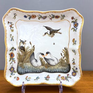 May include: A square porcelain plate with a scalloped edge. The plate is decorated with a floral border and a scene of two swans and a bird in flight. The swans are in a pond with reeds and the bird is flying above them.