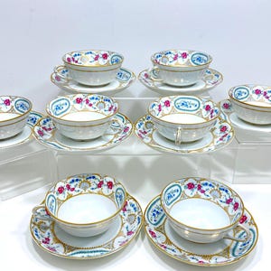 Könnte beinhalten: Sechsteiliges Set aus weißen Porzellan-Teetassen und Untertassen mit einem floralen Muster in Rosa-, Blau- und Goldtönen. Die Tassen und Untertassen haben ein zartes, verziertes Design.