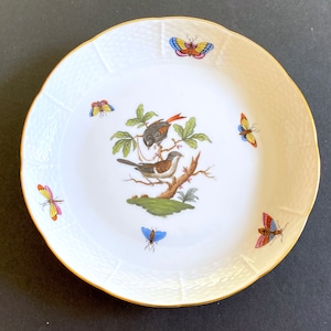 May include: A white porcelain plate with a gold rim features a hand-painted design of two birds perched on a branch with butterflies surrounding them. The plate has a textured surface.