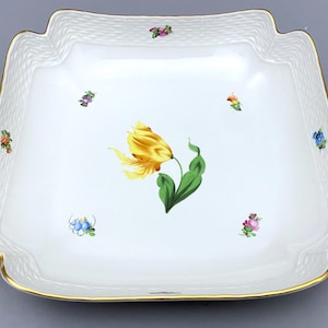 Könnte beinhalten: Eine quadratische Servierplatte aus weißem Porzellan mit einem erhabenen gewebten Muster am Rand. Die Platte zeigt eine gelbe Tulpe mit grünen Blättern und einer Streuung kleiner bunter Blumen.