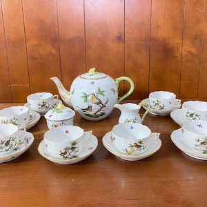 Puede incluir: Un juego de té de porcelana completo con tetera, cremera, azucarero y varias tazas y platillos. El juego presenta un motivo de pájaros y mariposas con ribetes dorados sobre un fondo blanco. La tetera tiene un asa verde.