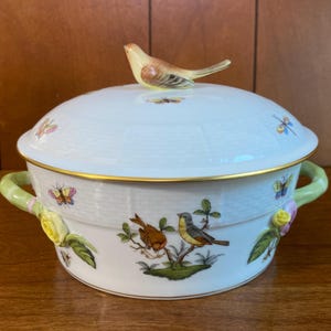 May include: A white ceramic serving dish with a lid, featuring a bird-shaped handle and painted bird and butterfly designs. The dish has green handles and gold trim, perfect for serving food.