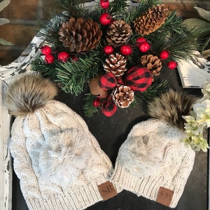 May include: Two white knit winter hats with faux fur pom poms. The hats have a cable knit pattern and a fluffy white pom pom on the top. The hats have a brown leather patch with the letters "SB" on them.