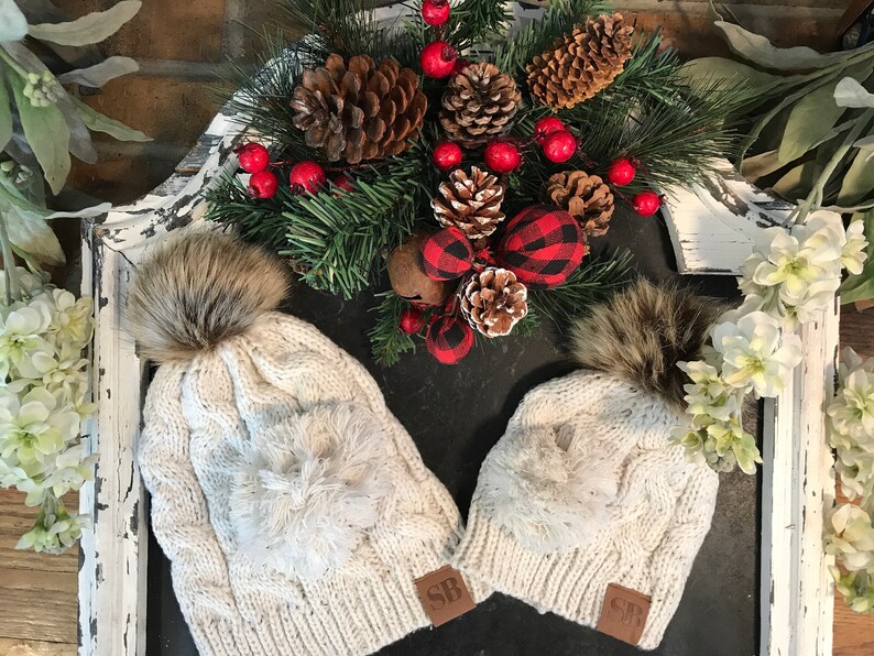 May include: Two white knit winter hats with faux fur pom poms. The hats have a cable knit pattern and a fluffy white pom pom on the top. The hats have a brown leather patch with the letters "SB" on them.