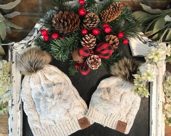 May include: Two white knit cable knit hats with faux fur pom poms. The hats have a small brown leather patch with the letters "SB" on them. The hats are sitting on a white wooden tray with a Christmas wreath in the background.