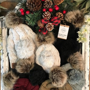 May include: Assortment of knit hats with faux fur pom poms in various colors, including white, black, gray, brown, and burgundy. The hats are displayed on a whitewashed wooden surface with a small brown tag that reads "Never a bad hair day".
