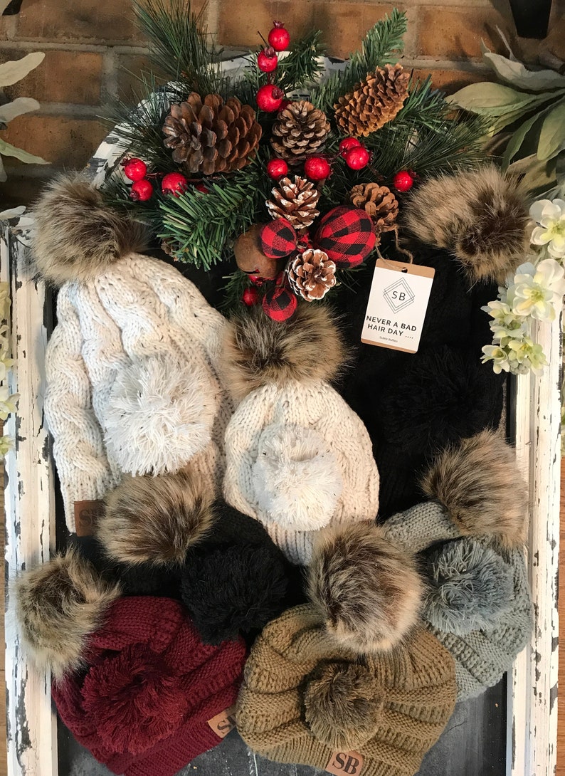 May include: Assortment of knit hats with faux fur pom poms in various colors, including white, black, gray, brown, and burgundy. The hats are displayed on a whitewashed wooden surface with a small brown tag that reads "Never a bad hair day".