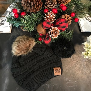 May include: Black knit beanie with a brown faux fur pom pom and a brown leather patch with the initials "SB".