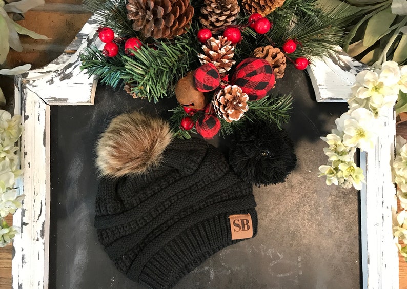 May include: Black knit beanie with a brown faux fur pom pom and a brown leather patch with the initials "SB".